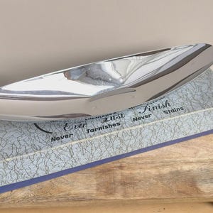 Op de afbeelding: Een gepolijste, zilverkleurige metalen serveerschaal met een slank, langwerpig ontwerp. De schaal rust op een doos met een patroon met de tekst "Never Lust, Ever Tarnishes, Never Finish Stains".