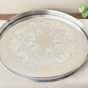 May include: A round silver-toned metal serving tray featuring a polished finish and an intricate embossed floral pattern in the centre. The tray has a raised gallery edge with a delicate pierced lattice design around the entire circumference.