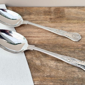 Op de afbeelding: Twee zilveren serveerlepels met sierlijke bloemen- en schelpmotieven op de handvatten. De lepels zijn hoogglans gepolijst en reflecteren het licht. Ze liggen op een houten oppervlak naast een witte doek.