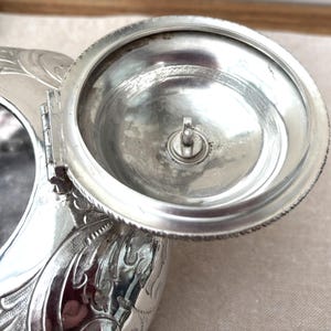 May include: A silver-toned metal inkwell featuring an ornate, embossed floral pattern on its exterior. The circular lid is attached by a hinge and is shown in an open position, revealing a central screw mechanism on the underside of the lid.