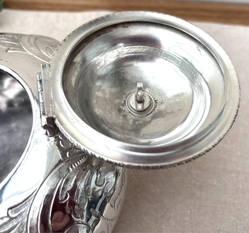 May include: A silver-toned metal inkwell featuring an ornate, embossed floral pattern on its exterior. The circular lid is attached by a hinge and is shown in an open position, revealing a central screw mechanism on the underside of the lid.