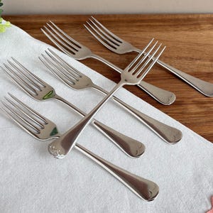 Op de afbeelding: Een set van vijf zilverkleurige metalen vorken in een waaiervormige opstelling. Elke vork heeft vier tanden en een gladde, afgeronde steel met een subtiel reliëfdetail aan de basis. De vorken zijn voorzien van een gepolijste afwerking.