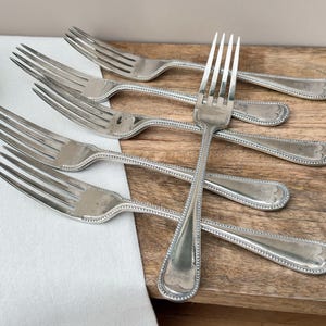 May include: Six silver forks with a beaded edge design are arranged on a wooden surface. The forks have four tines and a decorative handle. The background includes a white cloth and greenery.