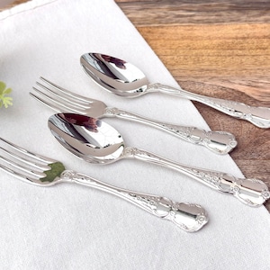 May include: Four pieces of silver flatware, including two forks and two spoons, are arranged on a white cloth. The silverware has ornate detailing on the handles and reflects the light. The background is a wooden surface.