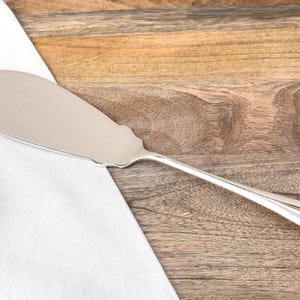 Puede incluir: Un servidor de pastel plateado con un borde festoneado y un mango largo y curvo. El servidor descansa sobre una superficie de madera junto a un paño blanco. Diseñado para servir pasteles.