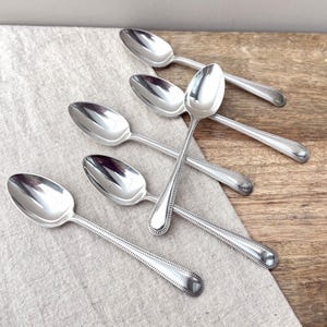 May include: A set of five silver-toned metal spoons with a polished finish. Each spoon features a slender handle decorated with a delicate beaded border along the edges and a rounded, smooth bowl, arranged in a layered pattern on a textured surface.