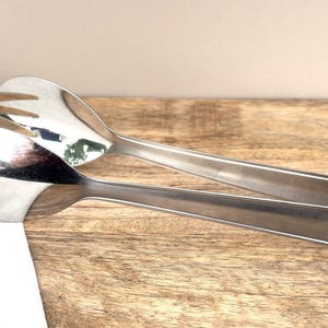 Peut inclure: Une paire de pinces de service brillantes en acier inoxydable. Une extrémité a la forme d'une cuillère, l'autre d'une fourchette. Les pinces reposent sur une surface en bois.