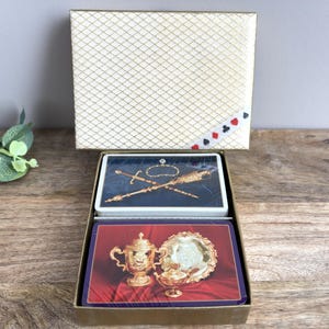 Könnte beinhalten: Ein Vintage-Spielkartenset in einer goldfarbenen Box mit einem Rautenmuster. Die Karten zeigen goldene und rote Bilder, darunter eine Trophäe, ein Schwert und ein Zepter. Die Box hat ein Band mit Spielkarten-Symbolen.