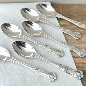 May include: Six silver spoons with decorative handles are displayed on a white linen cloth, set against a wooden surface. The spoons have a polished, reflective finish, and the handles feature a detailed floral design. The spoons are likely for serving or eating.