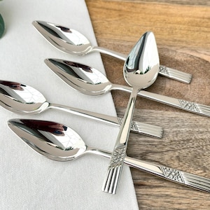 May include: A set of five silver spoons with a reflective surface. The spoons have a teardrop-shaped bowl and a handle with a geometric design. The spoons are arranged on a white cloth and a wooden surface.