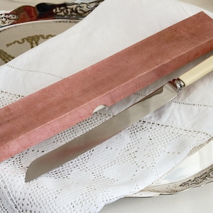 May include: A vintage cake knife with a cream-colored handle and a silver blade, next to a pink rectangular box. The knife and box rest on a white lace doily and a silver serving tray.