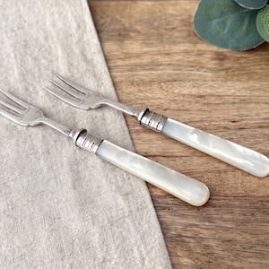 Op de afbeelding: Twee zilverkleurige metalen vorken met drie tanden en gladde, gebroken witte parelmoerachtige handvatten. Elke vork heeft een decoratieve metalen kraag met horizontale ribbels die de metalen kop verbindt met het rechthoekige, gepolijste handvat.