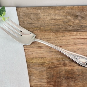 May include: A silver serving fork with four tines, on a wooden surface. The handle has an ornate floral design. The fork is on a white linen napkin, with greenery in the upper left. The fork's length is approximately 20 cm.