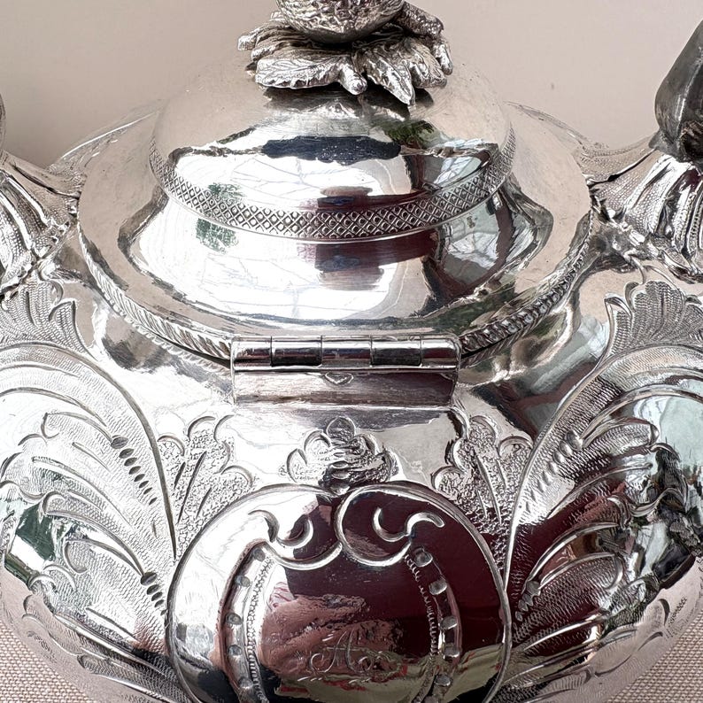 May include: A polished silver teapot featuring intricate embossed floral and leaf patterns across its surface. The lid is topped with a detailed fruit-shaped finial and a textured band. A central circular cartouche displays an engraved letter A.