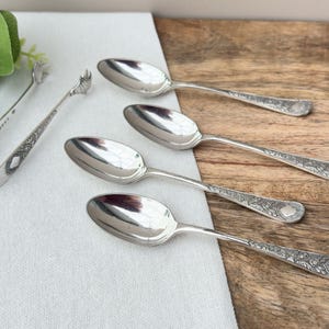 May include: A collection of silver serving spoons and a sugar tong, arranged on a wooden surface. The spoons feature detailed handles. The sugar tong is open and has a decorative design. Green leaves are in the background.