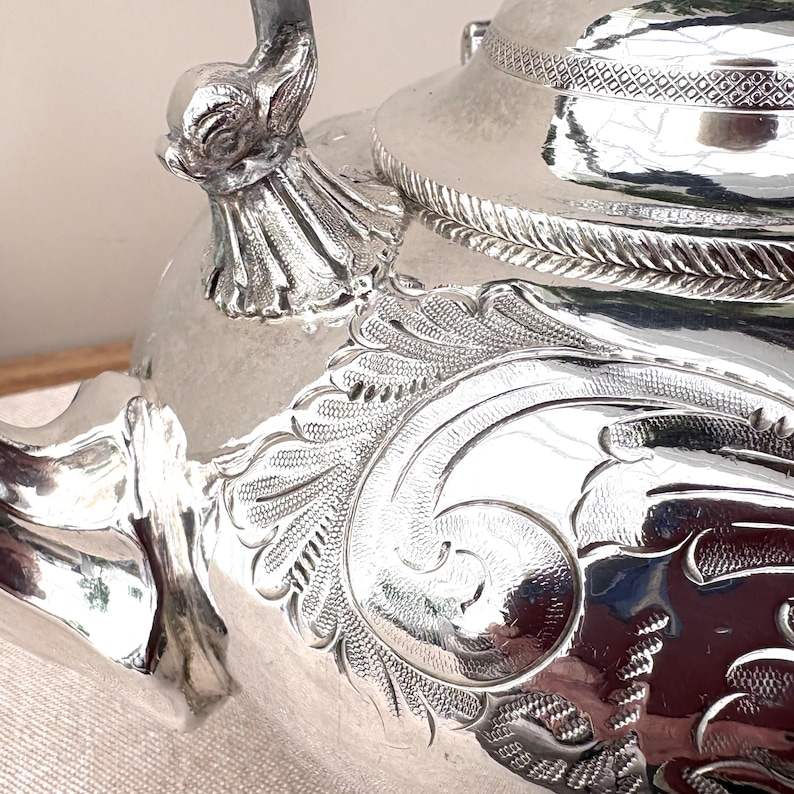 May include: A polished silver teapot featuring intricate engraved scrollwork and floral patterns on its body. The lid displays a textured border, while the handle attachment is sculpted into a detailed animal head shape with fluted metal accents.