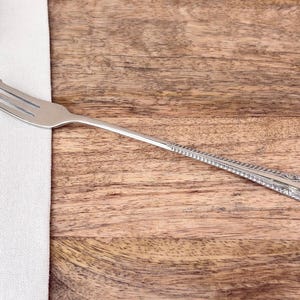 May include: A silver fork with three prongs, lying on a white cloth and a wooden surface. The fork has a decorative handle with a detailed pattern. The image also includes greenery.