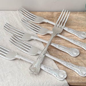 Op de afbeelding: Een set van vijf zilverkleurige metalen vorken in een waaiervormige opstelling. Elke vork heeft een gepolijste afwerking met een decoratief schelpmotief in reliëf aan de onderkant van het handvat. De tanden zijn slank en puntig.