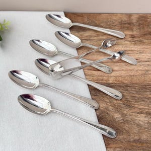 Può includere: Una collezione di cucchiai d'argento con coppette ovali e bordi perlati, disposti su un panno bianco e una superficie di legno. I cucchiai sono riflettenti e la disposizione suggerisce un'estetica vintage o antica.