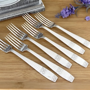 May include: Six silver forks with decorative handles are arranged diagonally on a wooden surface. The forks have tines and detailed patterns on the handles. A decorative mirror and purple flowers are in the background.