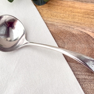 Peut inclure: Une louche en argent avec un bol arrondi et un long manche incurvé. La poignée porte une initiale gravée. La louche repose sur un tissu blanc et une surface en bois.