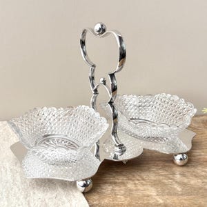 May include: A silver-toned metal serving stand featuring a central vertical handle and two clear glass bowls with a diamond-cut pattern. The base is a reflective silver-toned tray supported by small spherical feet, holding the two removable glass dishes.