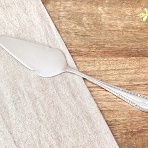 May include: A silver-toned metal cake server featuring a triangular blade with a scalloped edge and a slender handle decorated with subtle, raised linear detailing along the edges. The utensil has a smooth, polished finish throughout its surface.