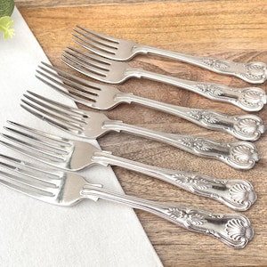 May include: Six silver forks with ornate floral and shell designs on the handles are arranged on a wooden surface. The forks are placed on a white cloth, with greenery in the upper left corner. The forks are a classic style.