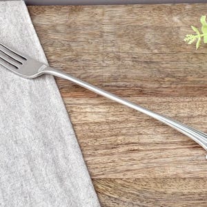 Puede incluir: Un tenedor de metal plateado con cuatro dientes y un mango largo y delgado. El mango termina en un remate decorativo en forma de abanico con un pequeño detalle de cuenta redondeada en el extremo, dando un acabado distintivo al utensilio.