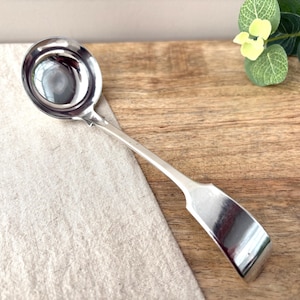 Puede incluir: Un cucharón de metal plateado pulido con un cuenco circular profundo y un mango largo y delgado que se ensancha en un extremo plano y curvado. El utensilio tiene una superficie lisa y reflectante, mostrando un diseño clásico para servir.