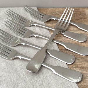 May include: A set of five silver-toned metal forks with four tines each. The forks feature a smooth, polished finish with a simple, flat handle design that widens slightly at the end. The utensils are arranged in a layered, overlapping pattern.