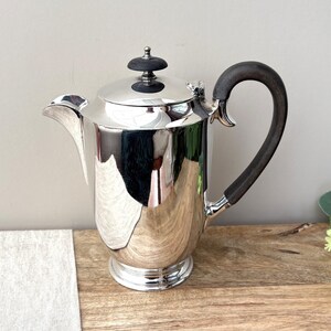 May include: A polished silver coffee pot featuring a curved spout, a circular base, and a dark brown wooden handle. The lid is topped with a small, round, dark brown wooden finial. The reflective surface shows distorted surroundings.