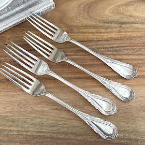May include: Four silver forks with decorative handles are arranged diagonally on a wooden surface. The forks have tines and ornate detailing on the handles. The background is a warm-toned wood.
