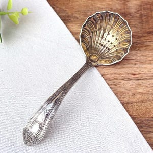 Puede incluir: Un colador de té de plata vintage con un cuenco en forma de concha y un mango largo y ornamentado. El cuenco tiene pequeños agujeros para colar las hojas de té. El mango presenta grabados detallados y un diseño ovalado. El colador descansa sobre un paño blanco y una superficie de madera.