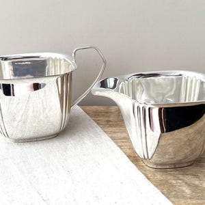 May include: A silver-toned sugar bowl and creamer set featuring a polished, reflective finish with vertical fluted detailing. The sugar bowl has two angular handles, while the creamer includes a single handle and a distinct pouring spout.