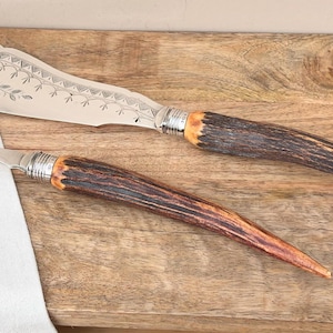 May include: A silver serving fork and a butter knife with decorative floral etching and antler handles. The utensils are displayed on a wooden surface, with a white cloth and greenery in the background.