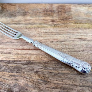 Op de afbeelding: Een zilveren vork met vier tanden en een decoratief handvat. De handgreep heeft sierlijke bloemen- en schelpmotieven. Het bestek is gemaakt van glanzend, reflecterend metaal en ligt op een houten oppervlak.