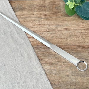 Può includere: Un tagliacarte in metallo color argento con una lama lunga e affusolata e un'impugnatura ad anello circolare alla base. L'utensile presenta una finitura liscia e lucida con una punta affilata, progettata per aprire buste di carta.