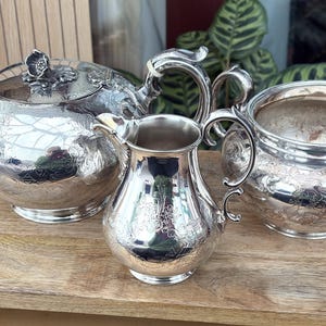 Può includere: Un servizio da tè in argento, composto da una teiera con coperchio floreale, una lattiera e una zuccheriera. Il set è esposto su una superficie di legno. I pezzi presentano dettagli ornati e una finitura riflettente.