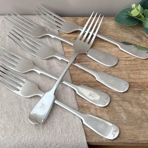 May include: A set of five silver-toned metal forks arranged in a fan pattern. Each fork features four tines and a smooth, rounded handle with a small, engraved monogram letter visible on the base of the handle. The forks have a polished finish.
