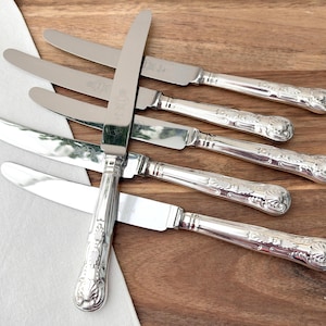 Könnte beinhalten: Sechs silberfarbene Messer mit verzierten Griffen liegen auf einer Holzoberfläche. Die glatten Klingen reflektieren das Licht, während die Griffe ein detailliertes Blumenmuster aufweisen. Ein weißes Tuch befindet sich unten links.