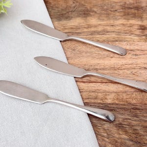 Può includere: Tre coltelli da burro argentati disposti su una superficie di legno e un panno di lino bianco. I coltelli hanno un design semplice con una lama arrotondata e un manico sottile. La superficie in legno ha un tono caldo e naturale.
