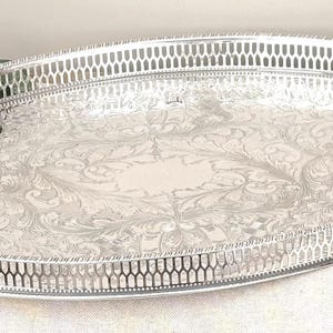 Könnte beinhalten: Ein ovales silberfarbenes Serviertablett mit einem komplexen gravierten Blumenmuster auf der Basis und einer dekorativen durchbrochenen Galerieumrandung. Das Tablett hat eine polierte metallische Oberfläche und einen glatten Mittelbereich.