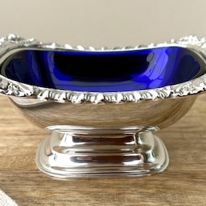 May include: A silver-toned pedestal bowl featuring a deep cobalt blue glass insert. The bowl has a rectangular shape with rounded corners and a decorative, textured rim. It stands on a sturdy, smooth, rectangular base with a polished finish.