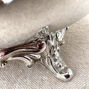 May include: A silver-toned bowl featuring a textured, matte-finish basin supported by a polished, ornate metal base. The base displays intricate, raised scrollwork and bead detailing, providing a decorative foundation for the curved vessel above.