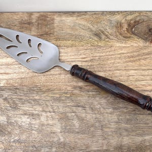 Puede incluir: Un servidor de pastel de acero inoxidable con un mango de madera marrón oscuro. El servidor tiene un diseño ranurado en la hoja. El servidor está sobre una superficie de madera.