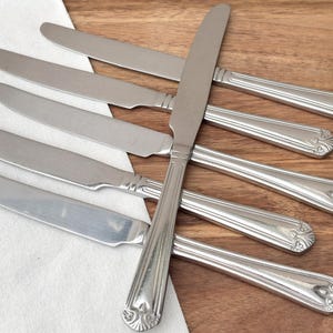 Puede incluir: Un juego de siete cuchillos de mesa de acero inoxidable dispuestos sobre una superficie de madera. Los cuchillos tienen un diseño de mango decorativo y se colocan sobre un paño blanco. Los cuchillos son para cenar.