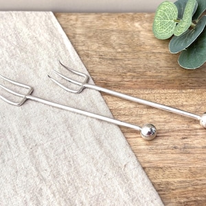 Puede incluir: Dos tenedores para aperitivo de metal en tono plateado, cada uno con una cabeza de tres puntas y un mango largo y delgado que termina en un pequeño remate esférico pulido. Los tenedores tienen un acabado liso y reflectante.