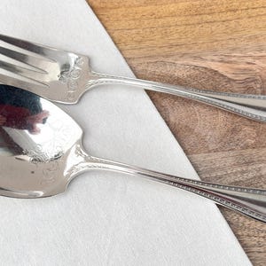 May include: Silver serving fork and spoon set. The fork has three prongs, and the spoon has a large, oval bowl. Both utensils have decorative floral patterns and beaded edges. The set is on a white cloth and a wooden surface.
