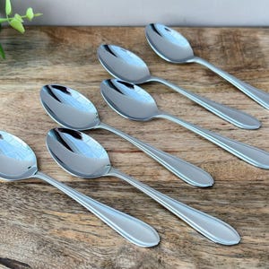 May include: Six stainless steel spoons with oval bowls and long, slender handles are arranged on a wooden surface. The spoons are reflective, and the wood grain is visible. A green plant is in the upper left corner.